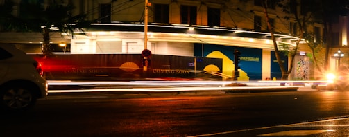 Nighttime urban scene with bright street lights creating light trails from passing cars. A partially illuminated building has signs indicating something is 'coming soon.' Trees are visible next to the road, and the atmosphere suggests a bustling city environment with a mix of artificial and natural elements.