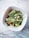 A fresh salad in a square white bowl on a marble surface, containing spinach leaves, grape tomatoes, crumbled feta cheese, black olives, and yellow bell pepper pieces.