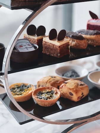 A stylish black metal stand holds an assortment of gourmet pastries. The top tier features elegantly decorated desserts including chocolate cakes and custard squares topped with chocolate discs. The lower tier presents savory quiches and muffins beside small bowls containing spreads.