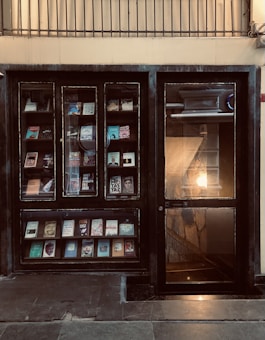 A storefront with glass windows displaying books on shelves. The book covers are of various colors and designs. There is a glass door on the right side, illuminated by warm light from inside, revealing a staircase.