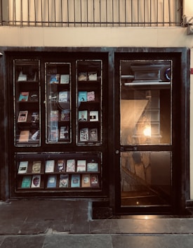 A storefront with glass windows displaying books on shelves. The book covers are of various colors and designs. There is a glass door on the right side, illuminated by warm light from inside, revealing a staircase.