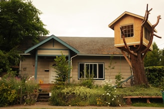 A quaint house surrounded by lush greenery and a wooden treehouse built on the branches of a large tree. The main structure is a single-story home with a sloped roof, a small porch, and several windows. The treehouse features multiple windows and is constructed from light wood, providing a whimsical contrast to the main house. The garden in front is thriving with a variety of plants and flowers.