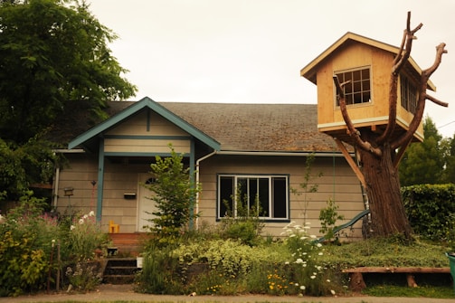 A quaint house surrounded by lush greenery and a wooden treehouse built on the branches of a large tree. The main structure is a single-story home with a sloped roof, a small porch, and several windows. The treehouse features multiple windows and is constructed from light wood, providing a whimsical contrast to the main house. The garden in front is thriving with a variety of plants and flowers.