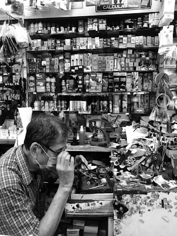 A small, cluttered workshop filled with various tools, boxes, and merchandise, including batteries, stationery, and small electronic items. A person wearing glasses and a face mask is focused on working at the station, using a tool on a small item. The walls are densely packed with shelves holding neatly arranged products.