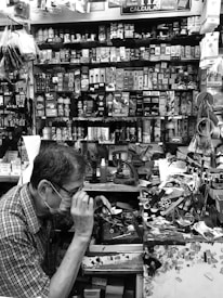 A small, cluttered workshop filled with various tools, boxes, and merchandise, including batteries, stationery, and small electronic items. A person wearing glasses and a face mask is focused on working at the station, using a tool on a small item. The walls are densely packed with shelves holding neatly arranged products.