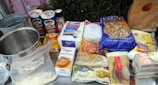 A variety of baking ingredients are arranged on a table, including Quaker Oats, Pillsbury flour, sugar, and a range of other items such as crackers, coconut, and barley. There is also a metal mixing bowl and a plastic ziplock bag containing some powder. Everything is neatly placed and clearly visible.