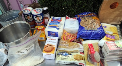 A variety of baking ingredients are arranged on a table, including Quaker Oats, Pillsbury flour, sugar, and a range of other items such as crackers, coconut, and barley. There is also a metal mixing bowl and a plastic ziplock bag containing some powder. Everything is neatly placed and clearly visible.
