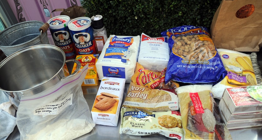A variety of baking ingredients are arranged on a table, including Quaker Oats, Pillsbury flour, sugar, and a range of other items such as crackers, coconut, and barley. There is also a metal mixing bowl and a plastic ziplock bag containing some powder. Everything is neatly placed and clearly visible.