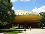 A large building with a distinctive dome shape, resembling a UFO, sits amidst lush greenery under a clear blue sky. People are walking around the area, and trees surround the structure. The foreground features a pathway leading toward the building.