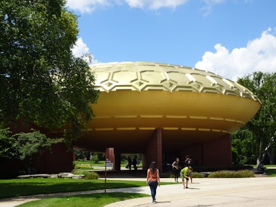 A large building with a distinctive dome shape, resembling a UFO, sits amidst lush greenery under a clear blue sky. People are walking around the area, and trees surround the structure. The foreground features a pathway leading toward the building.