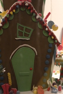 A whimsical gingerbread house decorated with colorful icing and candies. The structure features a slanted green door and a tilted window surrounded by various candy adornments. Brightly colored elements such as gumdrops and swirled lollipops accentuate the festive atmosphere.