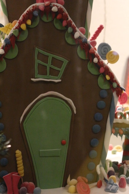 A whimsical gingerbread house decorated with colorful icing and candies. The structure features a slanted green door and a tilted window surrounded by various candy adornments. Brightly colored elements such as gumdrops and swirled lollipops accentuate the festive atmosphere.