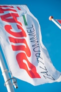 A close-up view of a white flag with bold red, green, and black text against a clear blue sky. The flag is attached to a pole with a metal fixture.