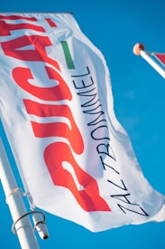 A close-up view of a white flag with bold red, green, and black text against a clear blue sky. The flag is attached to a pole with a metal fixture.