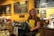 A cheerful barista stands behind the counter of a cozy café, wearing a green t-shirt and holding a receipt. The counter is adorned with a variety of baked goods under glass covers, and a credit card machine is visible. The background features several large blackboards with colorful chalk writing describing different menu items, along with various jars and a coffee machine.