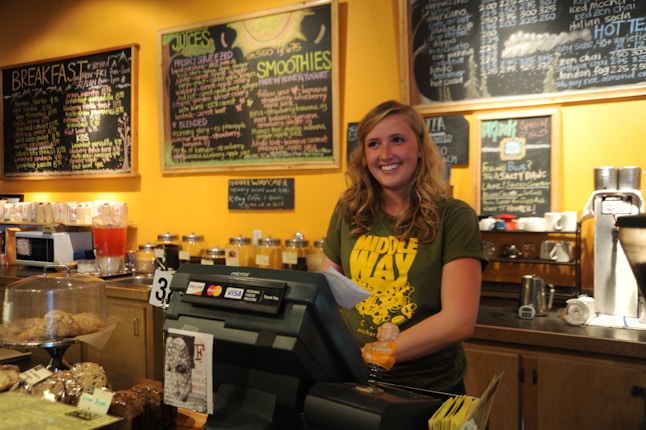 A cheerful barista stands behind the counter of a cozy caf&eacute;, wearing a green t-shirt and holding a receipt. The counter is adorned with a variety of baked goods under glass covers, and a credit card machine is visible. The background features several large blackboards with colorful chalk writing describing different menu items, along with various jars and a coffee machine.