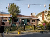 A small brick building with a traditional design features a teal door and several windows. In front of the building, there is a patio area with outdoor seating including chairs and tables. A black fence runs along the front. A woman is walking past, and a tree adds greenery to the scene. There are decorative strings of lights overhead.