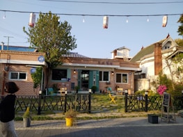 A small brick building with a traditional design features a teal door and several windows. In front of the building, there is a patio area with outdoor seating including chairs and tables. A black fence runs along the front. A woman is walking past, and a tree adds greenery to the scene. There are decorative strings of lights overhead.
