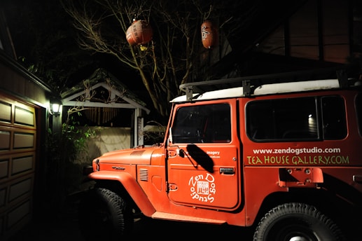 A red off-road vehicle is parked in a dimly lit area beside a wooden garage. The vehicle has branding for Zen Dog Studio on its side panels. Above the vehicle, there are two red lanterns hanging from tree branches, illuminating the scene with a warm glow. The environment suggests an artistic or rustic setting, possibly a gallery or studio space.