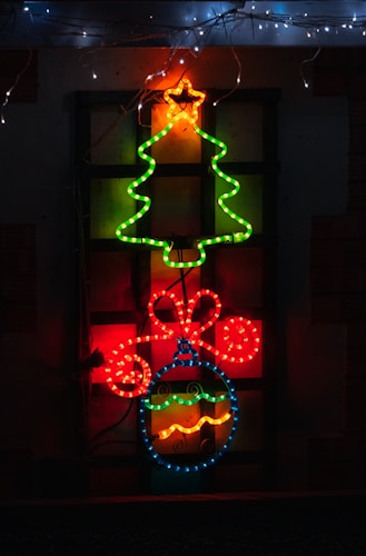 Bright, colorful Christmas lights form the shape of a Christmas tree with a star on top. Below the tree, a large ornament with ribbon is displayed, all set against a dark background.