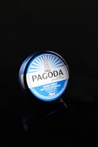 A round, blue tin container labeled 'Pagoda pastilles, Rasa Mint' is set against a black background. The design features a pagoda illustration and rays emanating from its top.
