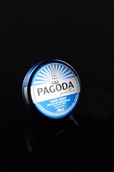 A round, blue tin container labeled 'Pagoda pastilles, Rasa Mint' is set against a black background. The design features a pagoda illustration and rays emanating from its top.
