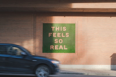 A wall artwork featuring the phrase 'THIS FEELS SO REAL' in large, prominent letters against a green background. The wall itself is made of brick, and a black car is moving past in the foreground.