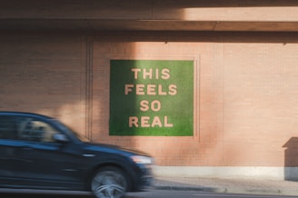 A wall artwork featuring the phrase 'THIS FEELS SO REAL' in large, prominent letters against a green background. The wall itself is made of brick, and a black car is moving past in the foreground.
