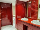 A modern bathroom interior with deep red cabinetry and tiles. The room features a double sink vanity with white basins and a tall mirror bordered by square red tiles. The cabinetry and countertops have a glossy finish, complementing the overall bold red color scheme. To the left, a closed, glossy red cabinet or door is visible. In the background, a glimpse of a white toilet can be seen.