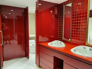 A modern bathroom interior with deep red cabinetry and tiles. The room features a double sink vanity with white basins and a tall mirror bordered by square red tiles. The cabinetry and countertops have a glossy finish, complementing the overall bold red color scheme. To the left, a closed, glossy red cabinet or door is visible. In the background, a glimpse of a white toilet can be seen.