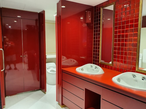 A modern bathroom interior with deep red cabinetry and tiles. The room features a double sink vanity with white basins and a tall mirror bordered by square red tiles. The cabinetry and countertops have a glossy finish, complementing the overall bold red color scheme. To the left, a closed, glossy red cabinet or door is visible. In the background, a glimpse of a white toilet can be seen.