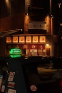 A dimly lit street scene featuring a taxi with a prominent green sign on its roof, parked in front of a warmly illuminated restaurant with wooden paneling and signage in Japanese characters.