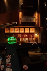 A dimly lit street scene featuring a taxi with a prominent green sign on its roof, parked in front of a warmly illuminated restaurant with wooden paneling and signage in Japanese characters.