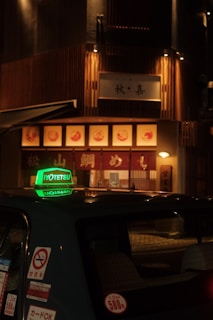 A dimly lit street scene featuring a taxi with a prominent green sign on its roof, parked in front of a warmly illuminated restaurant with wooden paneling and signage in Japanese characters.
