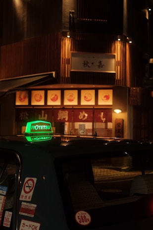 A dimly lit street scene featuring a taxi with a prominent green sign on its roof, parked in front of a warmly illuminated restaurant with wooden paneling and signage in Japanese characters.