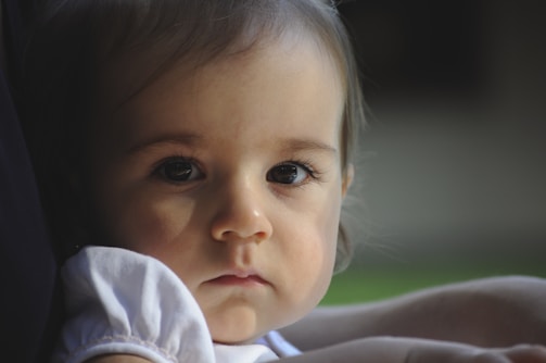 A close-up of a baby with wide, curious eyes and soft facial features. The baby's skin is soft and smooth, with faint shadows highlighting the contours of their face. They are wearing a white garment and are looking directly at the camera. The background is softly blurred, maintaining focus on the baby's expression.