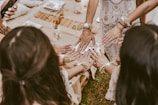 A group of women with their hands joined together, showing intricate henna designs and bracelets. The women are dressed in elegant attire, and the setting appears to be a festive or celebratory gathering. A table with decorative pieces is partially visible in the background.
