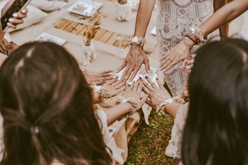 A group of women with their hands joined together, showing intricate henna designs and bracelets. The women are dressed in elegant attire, and the setting appears to be a festive or celebratory gathering. A table with decorative pieces is partially visible in the background.