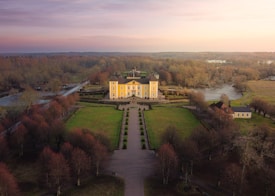 A large, yellow mansion is situated in the center of extensive manicured gardens, surrounded by a river and dense woodland. The mansion is of classic design with two tall towers and a symmetrical facade. A long driveway leads directly to the main entrance, flanked by trimmed trees and open green spaces. The surrounding landscape consists of bare trees, indicating a late autumn or early winter season. A bridge leading across the river can be seen to the left.