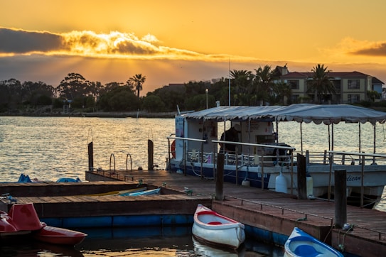 A serene waterfront scene at sunset with yellow and orange hues illuminating the clouds above. A small dock hosts several colorful kayaks, and a covered boat is moored next to it. Palm trees and a few buildings can be seen in the background, silhouetted against the sky.