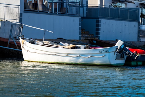 A white boat is docked along a calm body of water near a building. The boat has the registration number 'SR 1744 R' and is equipped with an outboard motor. The building in the background has a modern design with a balcony and metal railings.