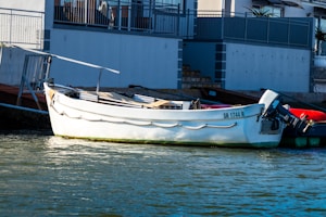 A white boat is docked along a calm body of water near a building. The boat has the registration number 'SR 1744 R' and is equipped with an outboard motor. The building in the background has a modern design with a balcony and metal railings.