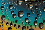 Numerous water droplets are scattered across a reflective surface, creating a rainbow-like effect with vibrant colors. The droplets magnify patterns in the background, adding a kaleidoscopic appearance.