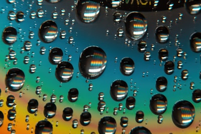Numerous water droplets are scattered across a reflective surface, creating a rainbow-like effect with vibrant colors. The droplets magnify patterns in the background, adding a kaleidoscopic appearance.