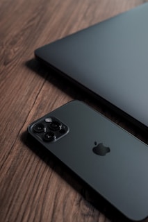 A sleek black smartphone and a partially visible laptop are placed on a wooden surface. The smartphone has a camera with multiple lenses and a distinctive logo.
