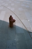 A red fire hydrant is positioned on a large tiled pavement. A metal rail or tube creates a curved line shadow across the tiles, adding a geometric element to the scene. The pavement consists of large rectangular tiles in varying shades of grey.