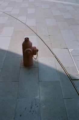 A red fire hydrant is positioned on a large tiled pavement. A metal rail or tube creates a curved line shadow across the tiles, adding a geometric element to the scene. The pavement consists of large rectangular tiles in varying shades of grey.