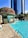 A luxurious outdoor swimming pool next to a modern skyscraper. The pool area features lounge chairs with umbrellas and a structure decorated with vibrant red and green foliage. Clear blue water contrasts with the light stone pool deck.