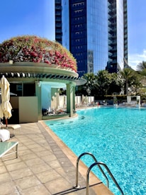 A luxurious outdoor swimming pool next to a modern skyscraper. The pool area features lounge chairs with umbrellas and a structure decorated with vibrant red and green foliage. Clear blue water contrasts with the light stone pool deck.