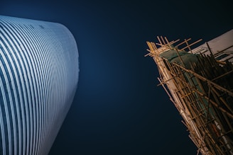 A modern skyscraper with a curved facade stands next to a building under construction covered in scaffolding. The scene is set against a deep blue evening sky, highlighting the contrast between the sleek, polished structure and the raw, unfinished construction.